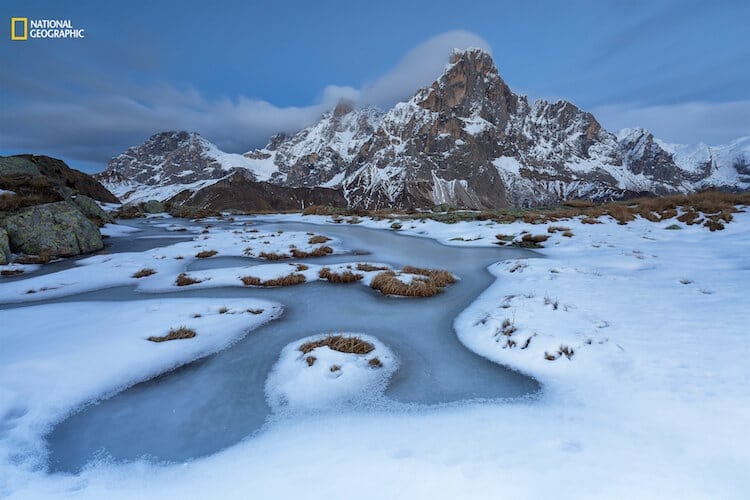 Alessandro Gruzza/2016 National Geographic Nature Photographer of the Year. Wild rink. 2nd Place—Landscape: The first cold days of winter have frozen the surface of a pond. The first snowfall has revealed its delicate beauty. A long shutter speed enhances the movement of the clouds around Mt. Cimon de la Pala, Paneveggio-Pale San Martino Natural Park, Italy