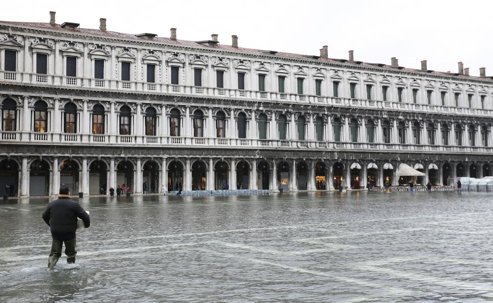 a man carries materials as he walks across the flooded st. mark's square during a period of seasonal high water and on the first day of carnival, in venice